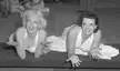 Marilyn Monroe and another woman in white polka-dot dresses lying on a dark floor, smiling in a pinup style.