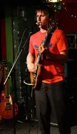 Lead guitar player in a red t-shirt singing into a microphone on an indoor stage
