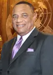 Perry Christie smiling in a grey suit, white shirt, and purple tie against a carved wooden wall