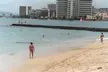 People in swimwear walking on a beach with a city skyline and high-rise buildings in the background