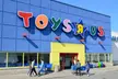 Group of people in casual fashion, including skinny blue denim jeans and white shirts, walking by a Toys "R" Us store.