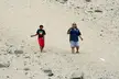 Two people, one with dark skin in a black t-shirt and red shorts, and one with fair skin in a blue shirt, walking on sand.