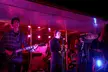 A 5-person band in streetwear, including a leather jacket and turtleneck, performing on a stage with pink and blue lights.