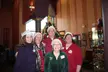 Five smiling friends in Santa hats, a green velvet jacket, and a plaid scarf posing by a Christmas tree.