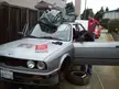 Two men in t-shirts and blue jeans working on a silver BMW car with a rollcage in a residential driveway