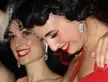 Two beautiful women with dark hair and red lipstick in burlesque costumes, featuring a red dress and black veil.