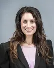 Leela Aheer, an Indian woman with long wavy brown hair wearing a black blazer and light purple v-neck blouse.