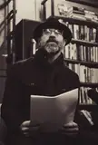 Alan Heathcock wearing a hat and glasses, holding paper in a library with bookshelves.