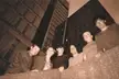 The band in 1994, a group of six people in jackets and shirts leaning over a concrete wall with a city skyline.