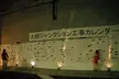 Two asian girls, one in a cardigan and short skirt, walking toward a construction calendar on a concrete wall at night