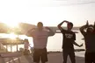 Three Asian girls in streetwear and hoodies making funny faces on a waterfront dock at sunset.