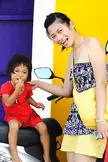 Woman in a floral strapless dress and yellow apron with a child in a red dress on a motorcycle