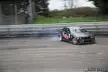Black BMW car drifting on a racing track with white smoke and a driver in a dark jacket
