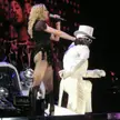 Blonde woman in a black sheer fringed outfit singing on stage with a man in a white suit and top hat.