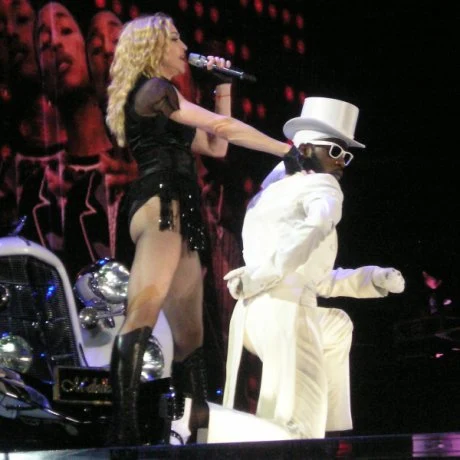 A faraway image of a female blond woman on stage in front of a white Speedster. The woman is wearing a white hat, black top and dancing. Two huge backdrops display two African American males in front of blinking lights. The one in the top backdrop looks towards the camera and smiles and the one in t