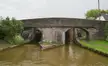 A brick bridge with two arches spanning a brown canal in Hassall, surrounded by greenery and a narrow path.