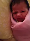 A person with dark hair and medium skin making a stink face while wrapped in a pink blanket on a yellow surface.