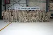 Group of 26 military personnel in desert camouflage uniforms posing in a hangar under a WE SUPPORT OUR TROOPS! banner