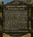Wooden historical marker for Peter Skene Ogden with a beaver carving, set against a landscape of grass and trees.