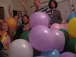 Six friends, including Asian girls, smiling and making funny faces behind a pile of colorful balloons.