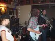 A four-person band with electric guitars and a drum set performing in a room with a dartboard.