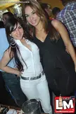 Two beautiful women in a group setting, one in a white ribbed top and hoop earrings, the other in a black dress and skirt.