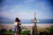 Asian girl in a blue sundress and brown shoulder bag taking a photo of the ocean by a wooden fence