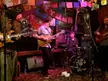 The Nice Ones band performing with electric guitars, bass guitar, and drum kit on a stage with colorful banners.