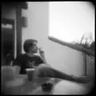 A person with short dark hair sitting barefoot on a balcony, smoking a cigarette during twilight.