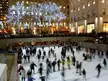 People in winter clothing ice skating at an outdoor city plaza rink with decorated trees and the Rock Center Cafe.