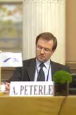 Lojze Peterle in a dark suit and glasses sitting at a table with a nameplate and a small houseplant.