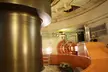A person in a yellow hard hat and dark clothing standing by orange industrial machinery and a spinning metallic cylinder