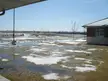 Spring weather landscape with melting snow, puddles, and a small building in a grassy field under a blue sky