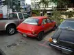 Red vintage car parked between a silver pickup truck and a black car on a Thailand city street with a palm tree.