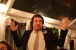 A man with curly brown hair in a black suit and purple flower boutonniere celebrating inside a white tent.