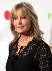 Bo Derek with blonde hair wearing a black lace v-neck top against a white background