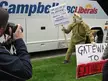 A person in a green dinosaur costume holding a protest sign by a campaign bus with a photographer nearby.