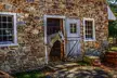 A white horse looking out from the open wooden door of a rustic stone building on a farm.