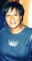 Sykes, an American voice actress, smiling in a blue t-shirt with a crown design and hoop earrings.
