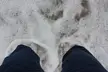A first-person view of legs in blue denim jeans standing in white sea foam on a beach.