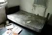 A dirty, worn-out mattress on a wooden bed frame in an abandoned trailer park room with peeling walls.