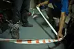 Two people in streetwear and sneakers handling red and white caution tape on a grey indoor floor.