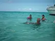 Four friends in swimwear swimming in a tropical turquoise ocean near a white boat.