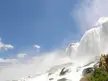 A group of five people in yellow raincoats standing beside a large waterfall cascading over rocks under a blue sky.