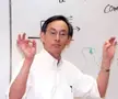 Steven Chu in a white shirt and glasses making OK signs in front of a whiteboard