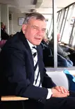 Dickie Bird smiling in a dark blue suit, white shirt, and striped tie, wearing a luxury watch