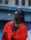 Ty Dolla Sign in an orange jacket and bandana sticking out his tongue with a cigar