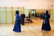 Kendo practitioners in blue armor and hakama using wooden swords in a gym with observers.