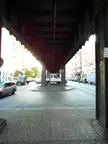 A person in dark clothing walking under the metal bridge at U-Bahnhof Kottbusser Tor in Berlin with cars and a white truck.