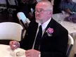 A man with a white beard and glasses wearing a black suit and luxury watch eating cake at a wedding reception jubilee.